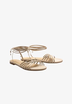 Beige flache Sandalen mit mehreren dünnen Riemen über den Zehen und Wickelbändern am Knöchel, verziert mit kleinen dekorativen Anhängern.