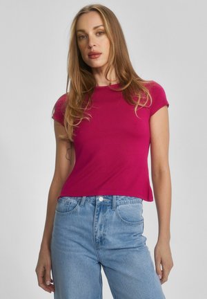 Młoda kobieta o długich blond włosach, ubrana w magentowy top z krótkim rękawem i jasnoniebieskie dżinsy z wysokim stanem, na jednolitym tle.