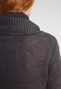 Dunkelgrauer gestrickter Pullover mit einem groben Kabelmuster, ausgestattet mit einem hohen Kragen und einem leicht lockeren Schnitt, der die Textur des Stoffes zur Geltung bringt.