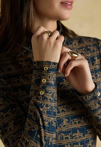 Kobieta nosząca wzorzystą granatową koszulę z złotymi guzikami oraz dwa duże błyszczące złote pierścionki na palcach w pobliżu twarzy i szyi.