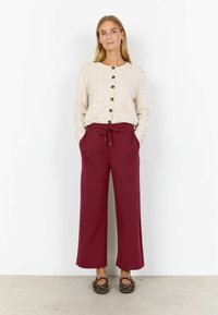 Cremfarbener Cardigan mit braunen Knöpfen, kombiniert mit bordeauxfarbenen weiteren Hosen mit Zugband und Taschen. Braune flache Schuhe mit Nieten.
