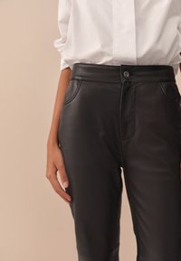 Pantalon en cuir noir à taille haute, présentant une texture lisse, des poches latérales et une fermeture simple par bouton en métal.