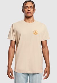 Beige bomulds t-shirt med korte ærmer og rund halsudskæring, med et orange grafisk tryk på venstre bryst.