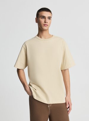 Giovane uomo in piedi con una mano in tasca, indossa una t-shirt beige oversize e pantaloni marroni su sfondo semplice.