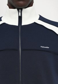 Marine blauwe zip-up jas met witte kraag en schouderaccenten, gemaakt van gestructureerde stof. Heeft een voorste rits en merklogo op de borst.