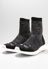 Schwarze gestrickte Socken mit strukturierter Oberfläche, hohem Knöcheldesign und weißen Katzen-Gesichtsmerkmalen auf den Zehen, ergänzt durch eine weiße Sohle.