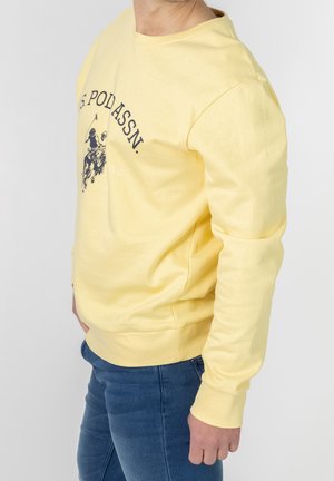 Persona che indossa una felpa gialla con una grafica di un giocatore di polo e del testo, abbinata a jeans blu, in piedi contro uno sfondo semplice.