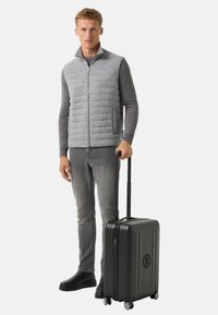 Graue Steppweste über einem grauen Langarmshirt, kombiniert mit grauen Jeans und schwarzen Stiefeln, während er einen schwarzen Hartschalenkoffer mit Rollen hält.