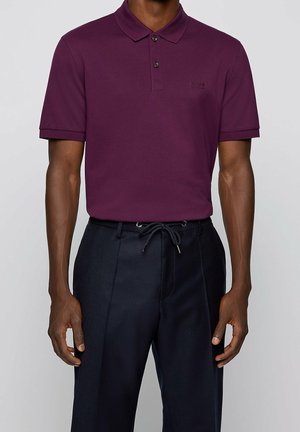 Homme portant un polo à manches courtes couleur prune rentré dans un pantalon noir à cordon de serrage, sur un fond uni.