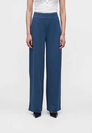 Femme portant un pantalon bleu taille haute à jambes larges avec poches latérales, associé à des escarpins noirs à bouts pointus et un haut blanc sans manches.