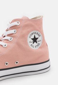 Zapatilla alta de lona rosa con cordones blancos, suela de goma negra y un parche circular de logo que presenta una estrella y el nombre de la marca.