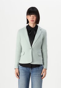 Svetle zelený blazer s texturovanou látkou, s klopami, zapínáním na knoflík a dvěma předními kapsami, v kombinaci s černou košilí a modrými džíny.