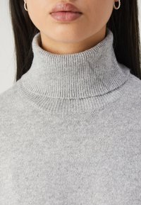 Femme portant un pull col roulé à côtes gris clair et des boucles d'oreilles en perles, lèvres et partie de la mâchoire visibles.