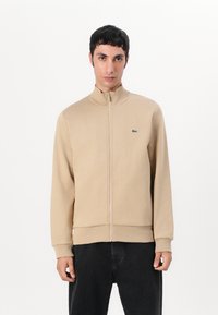 Beige zip-up jakke i et glatt materiale med høy krage, med en liten grønn logo og ribbede mansjetter og kant. Båret med mørke bukser.