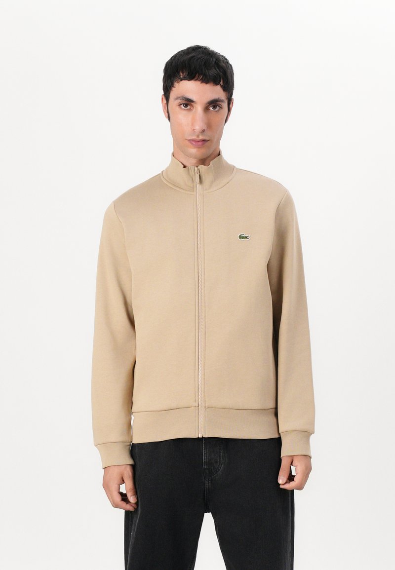Beige zip-up jakke i et glatt materiale med høy krage, med en liten grønn logo og ribbede mansjetter og kant. Båret med mørke bukser.