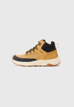 Tan and black hiking boot with padded ankle collar, metal eyelets, thick white sole, and brown laces on plain light background.