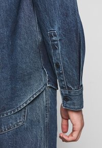 Camisa de denim en azul desgastado. Presenta un dobladillo curvado, puños abotonados y aberturas laterales. Tejido texturizado con costuras visibles y bolsillos.
