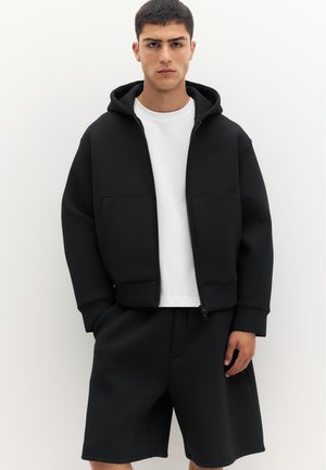 Jeune homme portant un sweat à capuche zippé noir sur un t-shirt blanc et un short noir assorti, debout devant un fond clair uni.