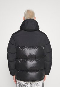 Svart pufferjacka med avtagbar huva, med en matt överdel och glansig underdel, quiltat mönster och elastiska ärmslut.