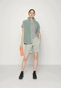 Kvinne står med hånden i lomma, iført grå t-skjorte, lysegrønn utenermet vest, matchende shorts, svarte sko, og holder en oransje vannflaske.