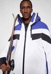 Lyžiarska bunda so bielym telom, modrými a čiernymi pruhmi, vysokým golierom a vreckami na zips. Vyrobená z materiálu odolného voči poveternostným vplyvom.