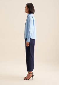 Camicia azzurro chiaro con maniche lunghe, abbinata a pantaloni navy a sigaretta e sandali neri con tacco alto, mostrata di profilo.