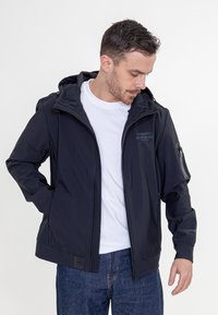 Zwarte waterdichte jas met een capuchon, voorzien van een ritszak op de mouw en elastische manchetten. Draagt over een wit T-shirt en denim jeans.