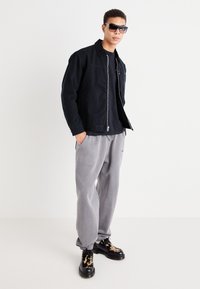 Svart bomullsjakke med glidelås foran, grå joggebukser og svarte sko med camouflage-mønster. Modell med mørke solbriller.