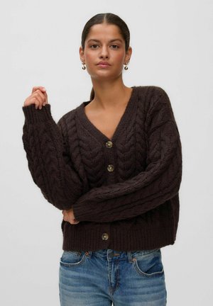 Vrouw met donker haar in een lage paardenstaart draagt een donkerbruine kabelgebreide button-down cardigan en blauwe spijkerbroek tegen een eentonige achtergrond.