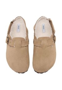 Ett par beigefärgade mocka slip-on klackar med justerbara sidostrapar och vit fluffig foder, sett ovanifrån på en vit bakgrund.