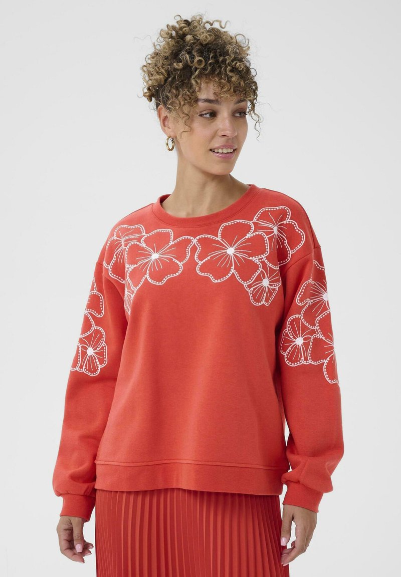 Femme aux cheveux bouclés portant un sweat-shirt rouge avec des motifs de fleurs blanches et une jupe plissée rouge assortie, regardant sur le côté.