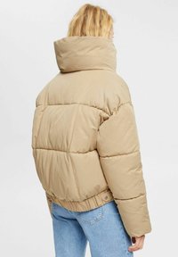 Chaqueta acolchada beige con cuello alto, tela texturizada y costuras horizontales. Cuenta con dobladillo elástico y costuras laterales visibles.