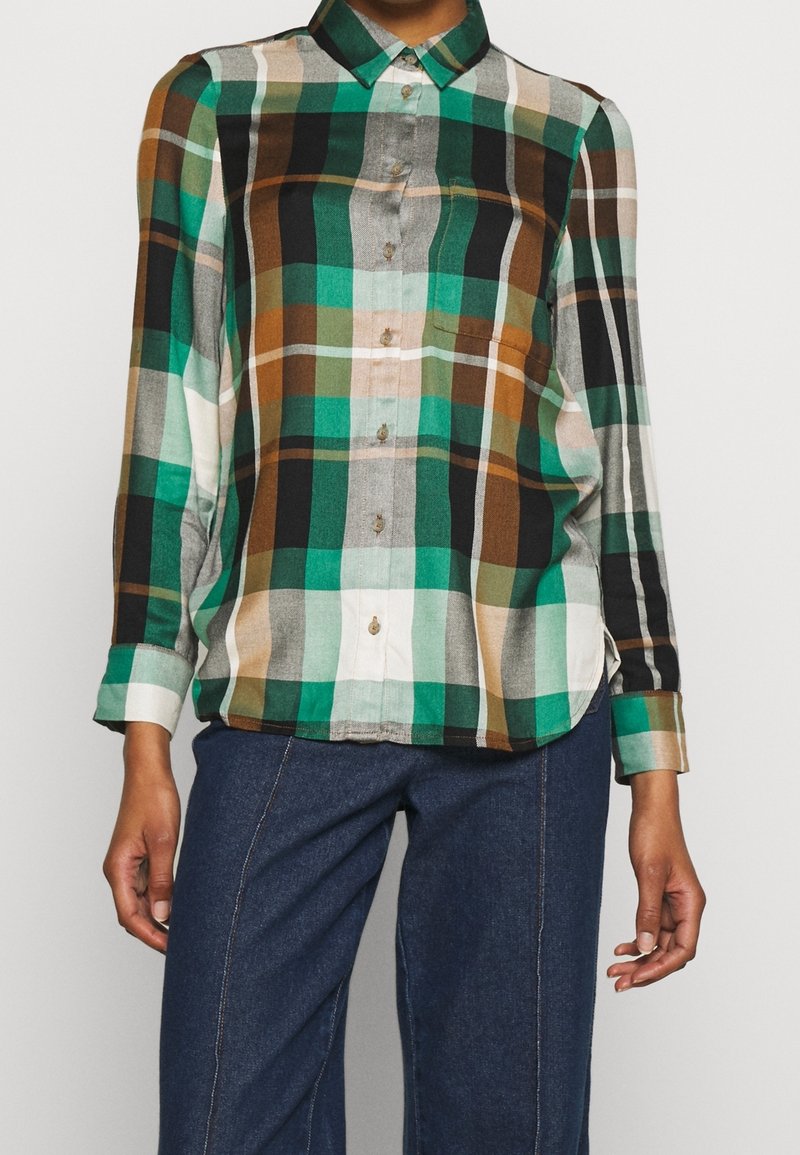 Chemise à manches longues boutonnée avec motif à carreaux vert, blanc, noir et marron, portée avec un pantalon bleu foncé.
