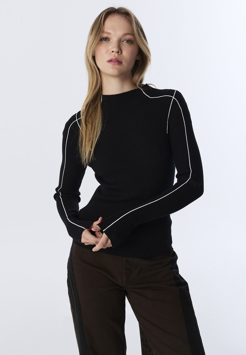 Femme avec de longs cheveux blonds portant un pull à manches longues côtelé noir avec des bordures blanches et un pantalon marron avec des rayures noires sur les côtés, sur fond blanc.