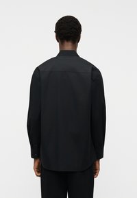 Άτομο με κοντά σγουρά μαλλιά που φοράει μακρυμάνικο μαύρο πουκάμισο και μαύρο παντελόνι, εμφανίζεται από πίσω σε απλό ανοιχτό φόντο.