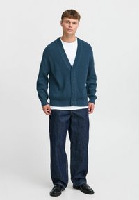 Mørkeblå ribbestrikket cardigan med dyp V-hals og fem knapper, båret over en hvit skjorte og kombinert med løse mørke denimjeans.