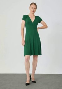 Rochie verde până la genunchi din material moale, cu design în formă de înfășurare, mâneci scurte și fustă evazată. Purtați cu pantofi cu toc negri pe un fundal deschis.