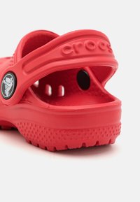 Red Crocs sandals made of foam, featuring ventilation holes, a rear strap, and textured soles for grip. Logo detail on the side.