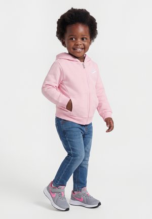 Lille barn smiler, iført en pink hættetrøje med lynlås, blå jeans og grå sneakers med pink detaljer, står mod en ensartet lys baggrund.