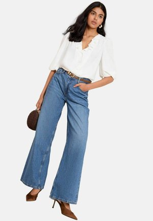 Jeune femme portant une blouse blanche à volants, un jean bleu taille haute à jambes larges, des talons marron, tenant un petit sac à main marron, posant avec une main dans la poche.