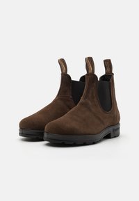 Botas Chelsea de ante marrón con paneles laterales elásticos, tiradores y suelas de goma negra. Textura suave y diseño de puntera redondeada.