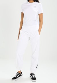 Osoba v bílém tričku s krátkým rukávem a bílých teplácích, s černobílými teniskami, stojící s rukama v kapsách.