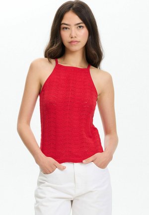 Mujer con cabello largo y oscuro, que lleva un top rojo de punto sin mangas y pantalones blancos, de pie con las manos en los bolsillos frente a un fondo blanco.