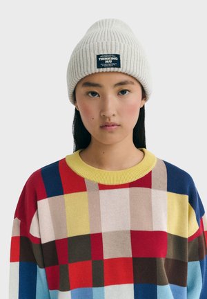 Junge Frau trägt eine cremefarbene Strickmütze mit einem Etikett und einen mehrfarbigen karierten Pullover mit gelbem Kragen vor einem schlichten Hintergrund.