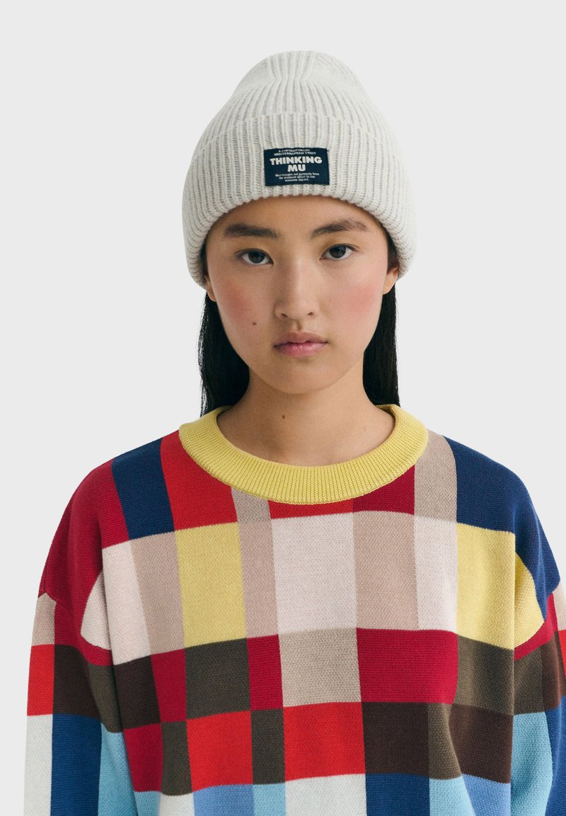 Unge kvinde iført en cremet strikket hue med et mærke og en flerfarvet ternet sweater med gul krave foran en ensfarvet baggrund.