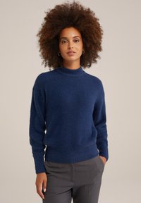 Marinblått texturerat tröja med hög krage och långa ärmar, tillverkat av mjukt material. Bärs över grå byxor med avslappnad passform.