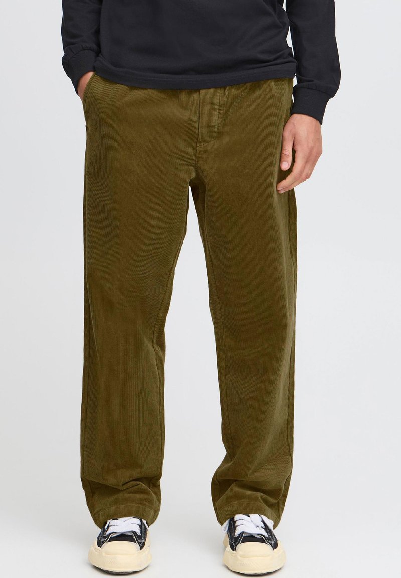 Person mit lockerer olivgrüner Cordhose, schwarzem langärmligem Shirt und schwarzen Sneakers mit weißen Sohlen, Hände in den Taschen.