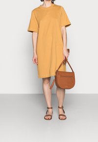 Persona que lleva un vestido de manga corta amarilla mostaza, sosteniendo un bolso de hombro de cuero marrón y usando sandalias marrones con tiras al tobillo.