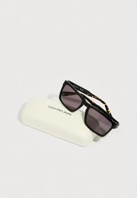 Czarne okulary przeciwsłoneczne z szarymi szkłami i zausznikami w wzór żółwia, spoczywające na białym etui oznaczonym napisem "Calvin Klein Jeans".