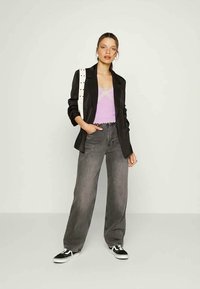 Jovem mulher de pé com uma mão no bolso, vestindo um blazer preto, uma camisola de renda lilás, jeans largos cinzentos, ténis pretos e uma mala de ombro branca.